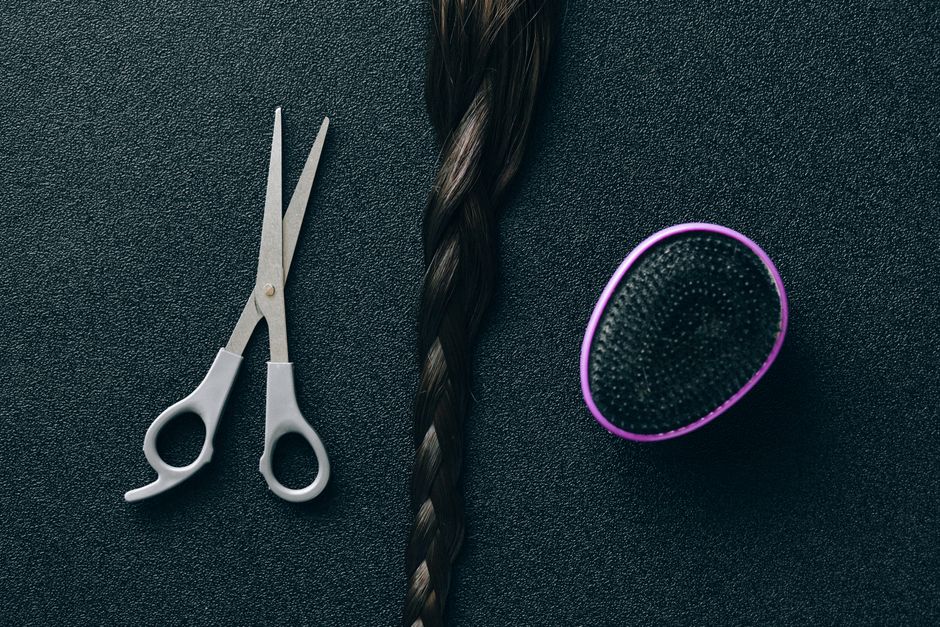Imagem de uma tesoura, uma mecha de cabelo cacheado e um aparelho de escovação térmica em fundo escuro, relacionada a cuidados capilares e beleza.
