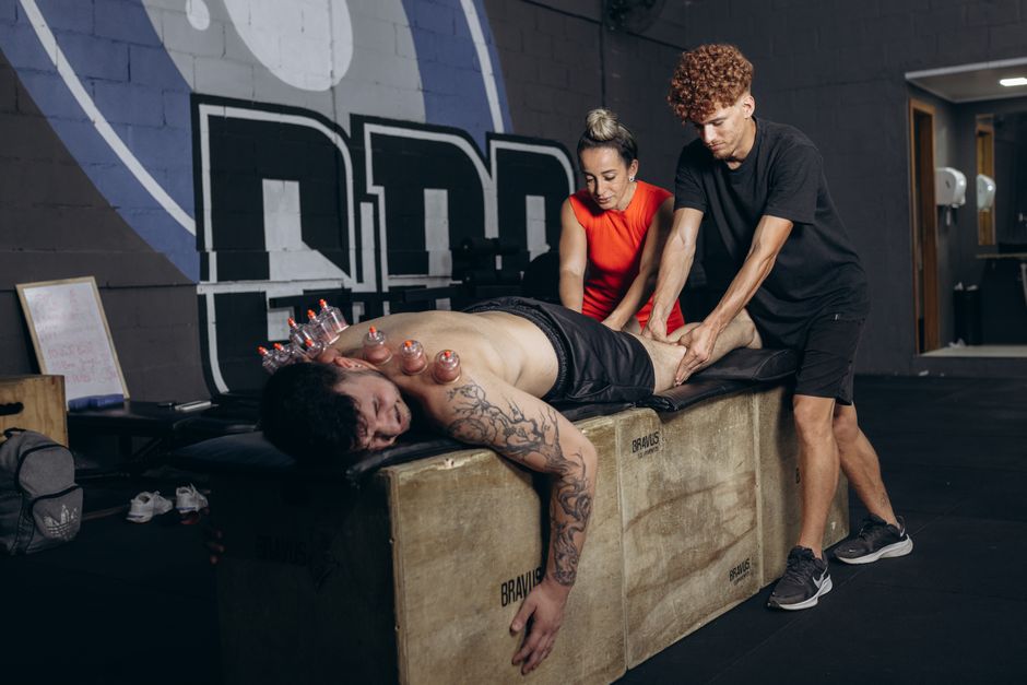 Imagem de uma sessão de preparação ou recuperação física, com duas pessoas auxiliando um homem que está deitado sobre um banco de exercícios, recebendo estímulos ou massagens manipulares, em um ambiente de academia ou treinamento funcional.