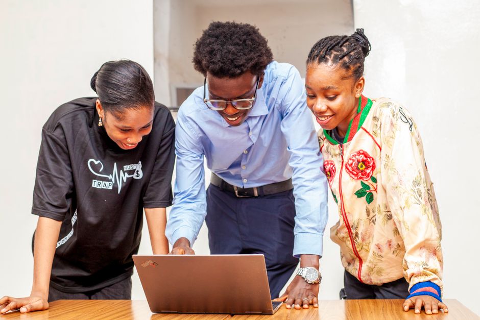 Três jovens entusiasmados colaborando em um projeto utilizando um laptop, refletindo inovação, trabalho em equipe e criatividade em um ambiente de trabalho cotidiano.