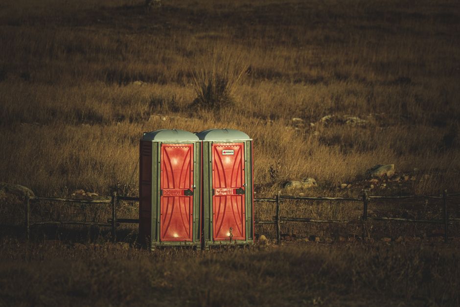 Imagem de dois banheiros portáteis vermelhos em meio a um campo rústico, ao entardecer, transmitindo uma sensação de isolamento ou preparação para eventos ao ar livre.