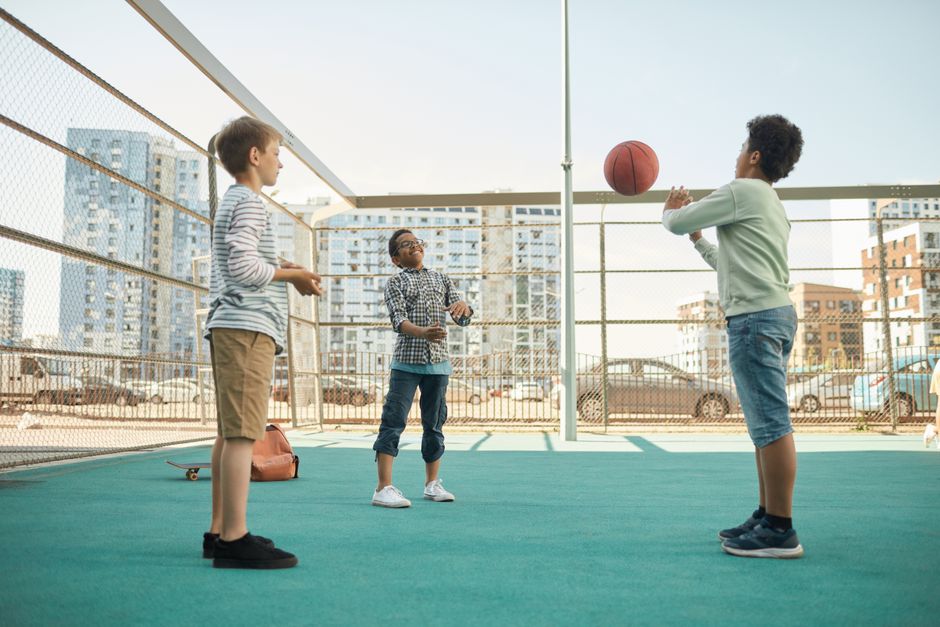 Crianças brincando de jogar basquete ao ar livre em uma quadra de rua, com edifícios residenciais ao fundo.