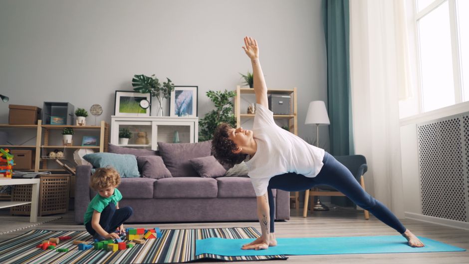 Mulher praticando yoga em casa enquanto uma criança brinca com blocos de montar ao seu lado, em um ambiente acolhedor e bem iluminado.