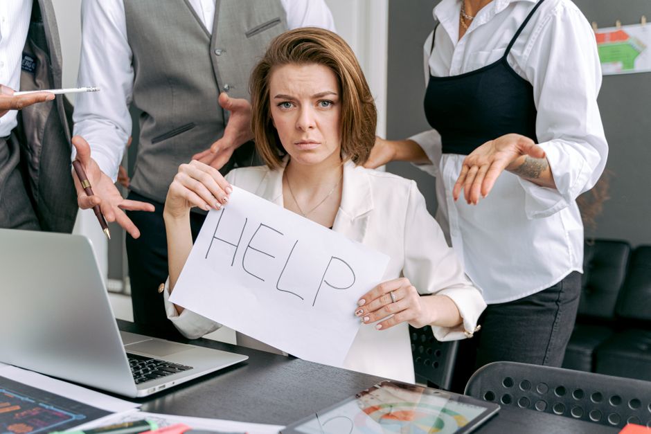 Mulher com expressão de preocupação segurando um cartaz com a palavra 'HELP', cercada por pessoas em um ambiente de trabalho, refletindo sobre dificuldades no trabalho ou na vida profissional.