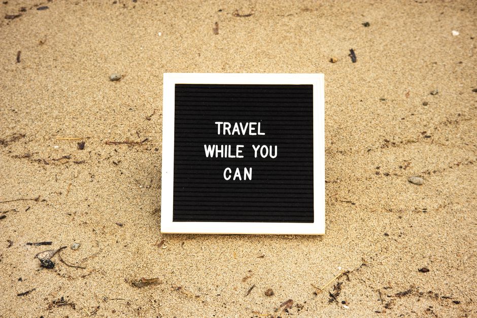Quadro com mensagem motivacional sobre viajar enquanto se pode, colocado na areia de uma praia.