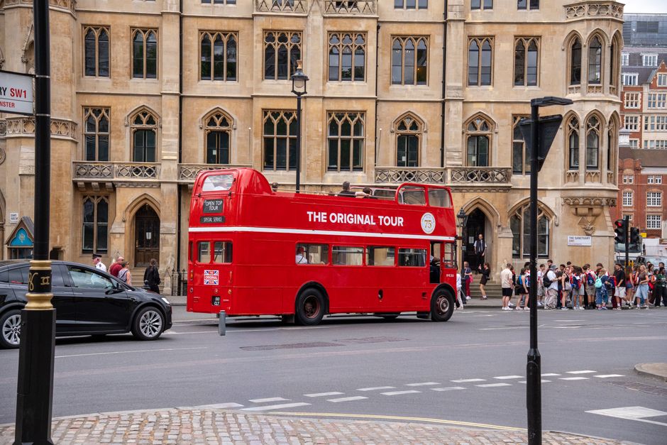 Imagem de um ônibus de turismo de dois andares vermelho em uma rua movimentada de uma cidade histórica com arquitetura antiga e turistas aguardando na calçada.