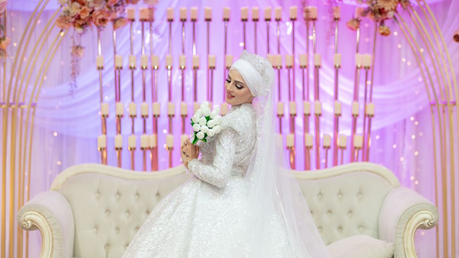 Noiva sorridente em um casamento, segurando um buquê de flores brancas, com um cenário decorado em tons de rosa e iluminação suave ao fundo.