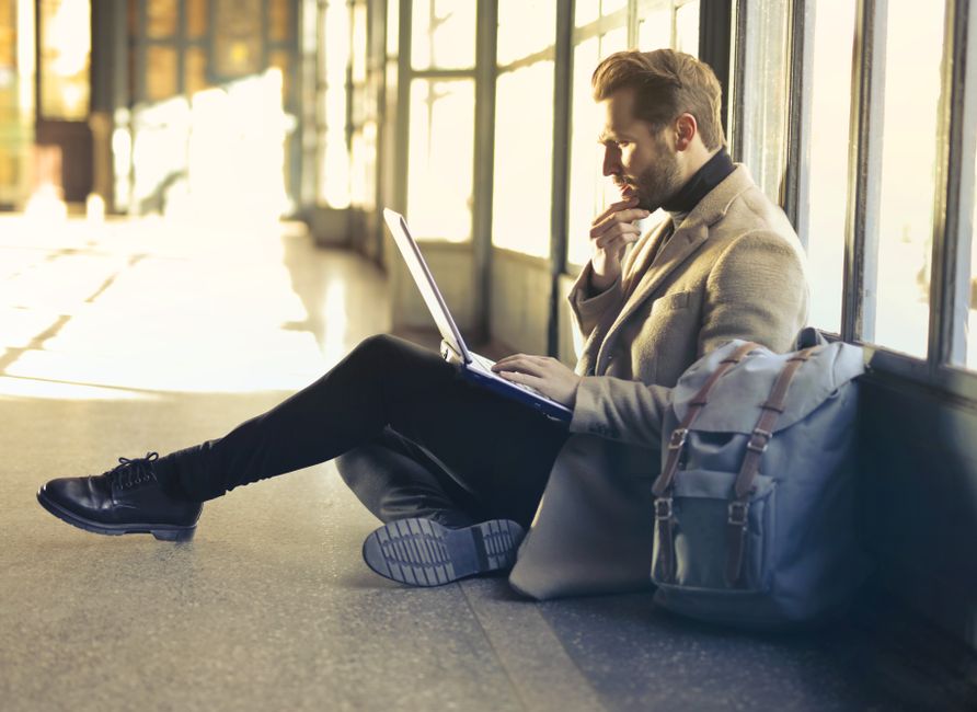 Homem pensativo usando laptop em um ambiente de espera com luz natural entrando pelas janelas, sugerindo reflexão ou pausa durante uma viagem ou pausa no trabalho.