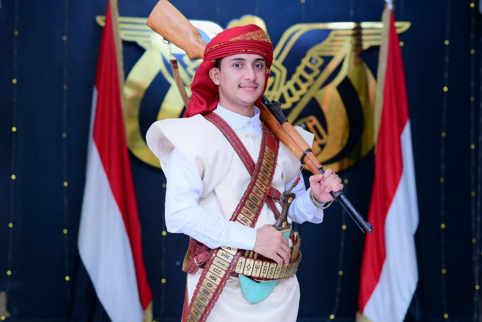 Homem vestido com traje tradicional e chapéu, segurando um rifle, em uma celebração cultural com bandeiras ao fundo.