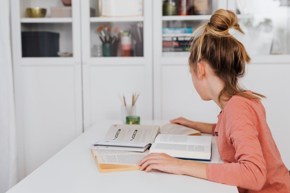 Jovem estudando em um ambiente doméstico com livros abertos, focada na leitura ou pesquisa.