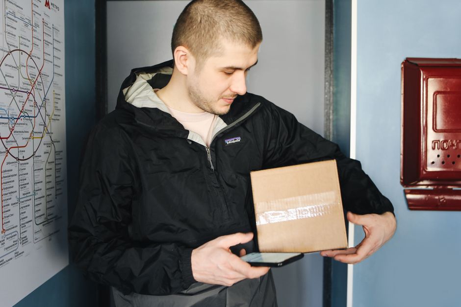 Homem aguardando na porta de um ponto de entrega, usando celular e segurando uma caixa de encomenda, simbolizando o processo de recebimento e logística de entregas.