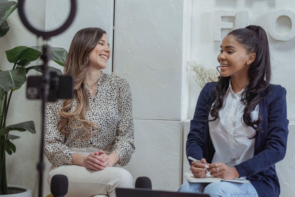 Duas mulheres em uma conversa descontraída durante uma entrevista ou sessão de gravação para blog ou podcast, em ambiente de escritório moderno.