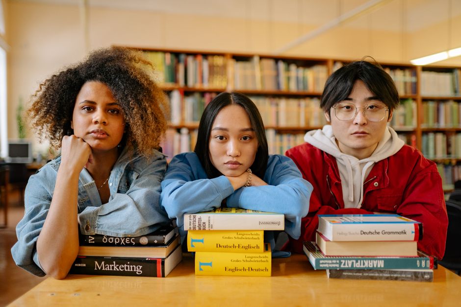 Três estudantes com expressão séria sentados com livros de idiomas e leitura em uma biblioteca universitária, representando estudo e aprendizagem.