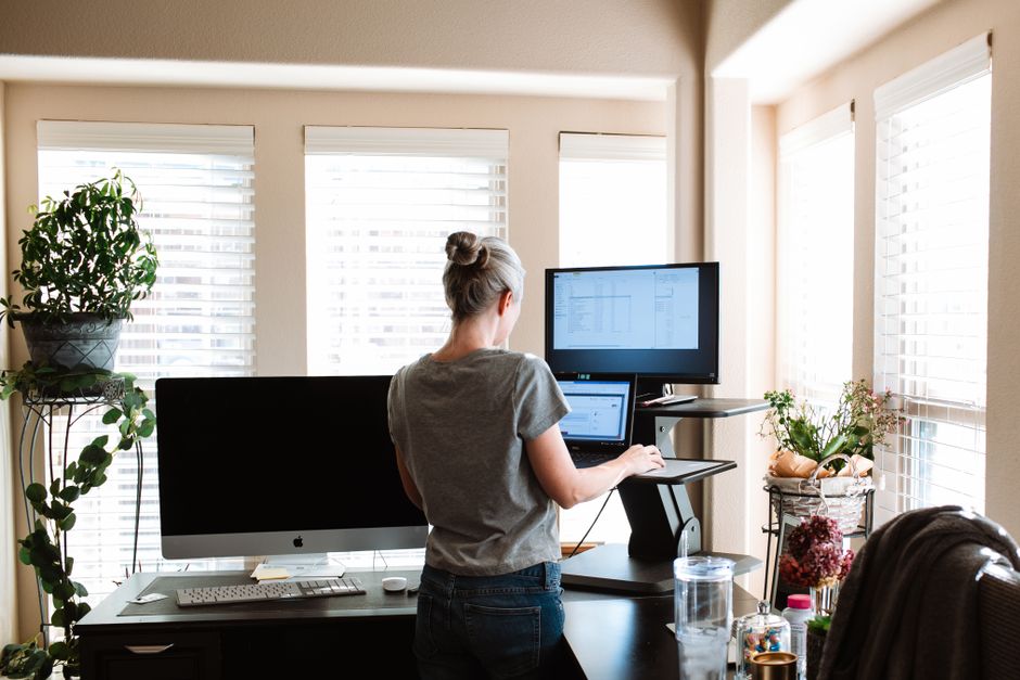 Mulher trabalhando em home office, usando computador e organizando seu espaço de trabalho com luz natural e plantas ao redor.