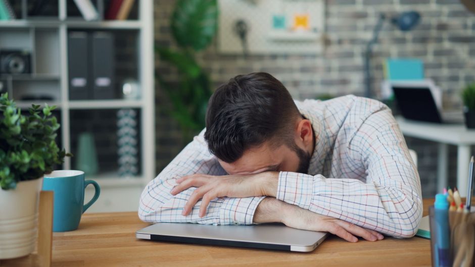 Homem com expressão de cansaço ou estresse, descansando a cabeça em um laptop em um ambiente de escritório moderno.