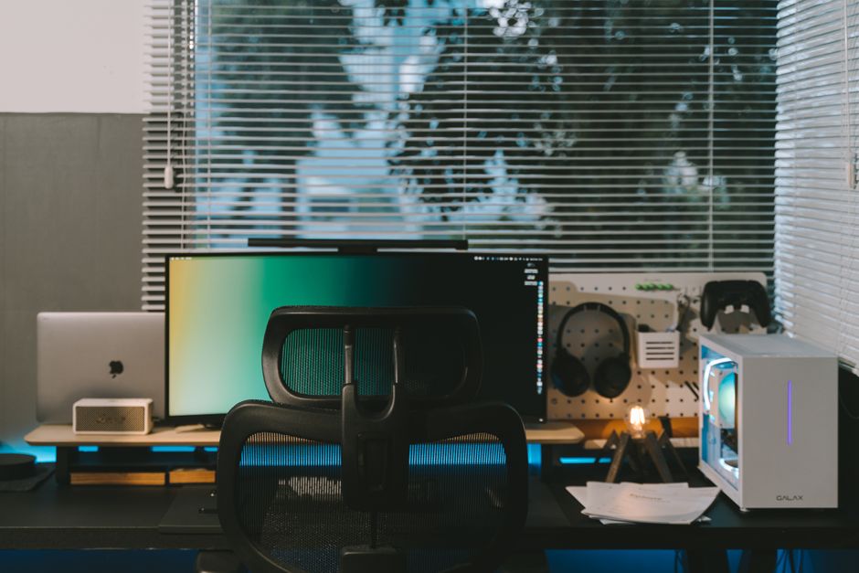 Imagem de um escritório com mesa de trabalho, monitor de computador, cadeira ergonômica, acessórios tecnológicos e janelas com persianas, ideal para conteúdo sobre home office ou produtividade.