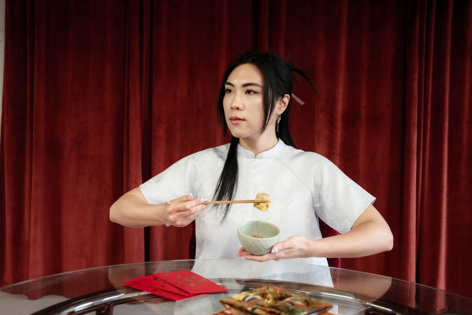 Mulher em traje tradicional asiático segurando um bolinho com pauzinhos em um ambiente com cortinas vermelhas, simbolizando cultura e gastronomia oriental.
