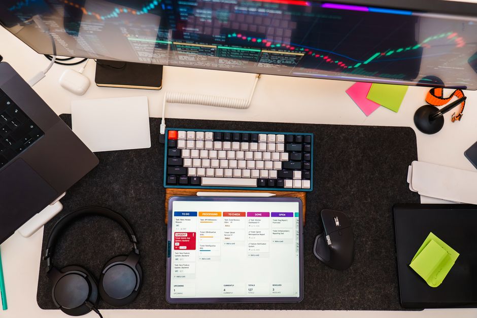Imagem de um espaço de trabalho moderno com foco em gestão de projetos ou produtividade, incluindo computador com gráficos, tablet com agenda, teclado mecânico, fones de ouvido e material de escritório.