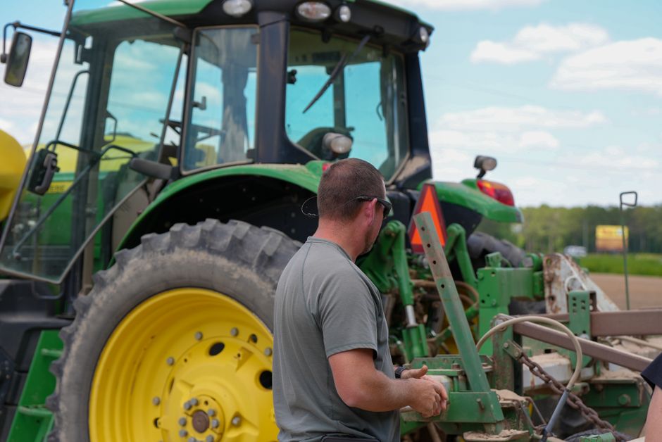 Homem observando e verificando equipamentos agrícolas em um campo com trator ao fundo, enfatizando o trabalho no setor agrícola.