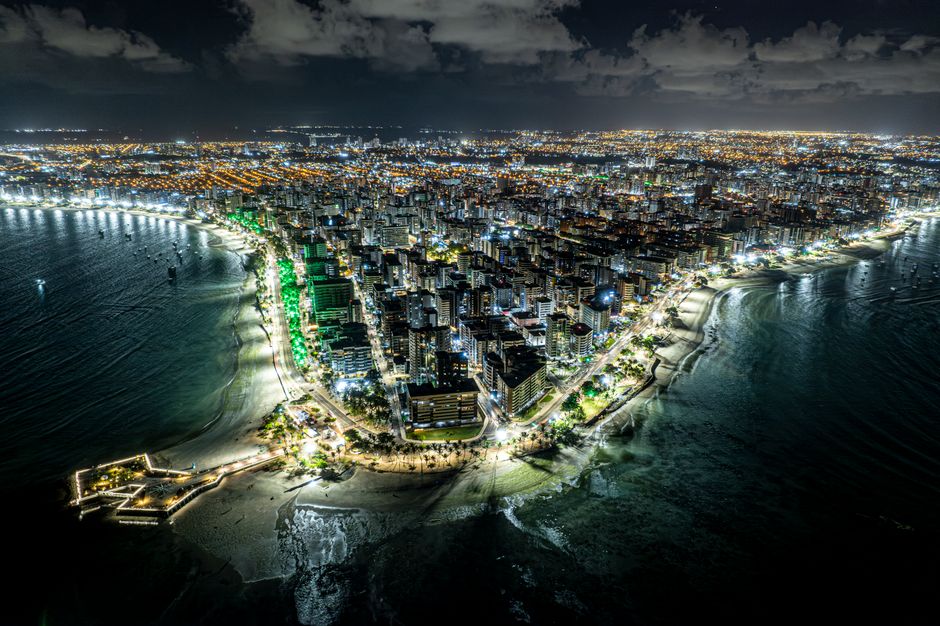 Vista panorâmica noturna de uma cidade costeira com iluminação urbana e praias na orla.