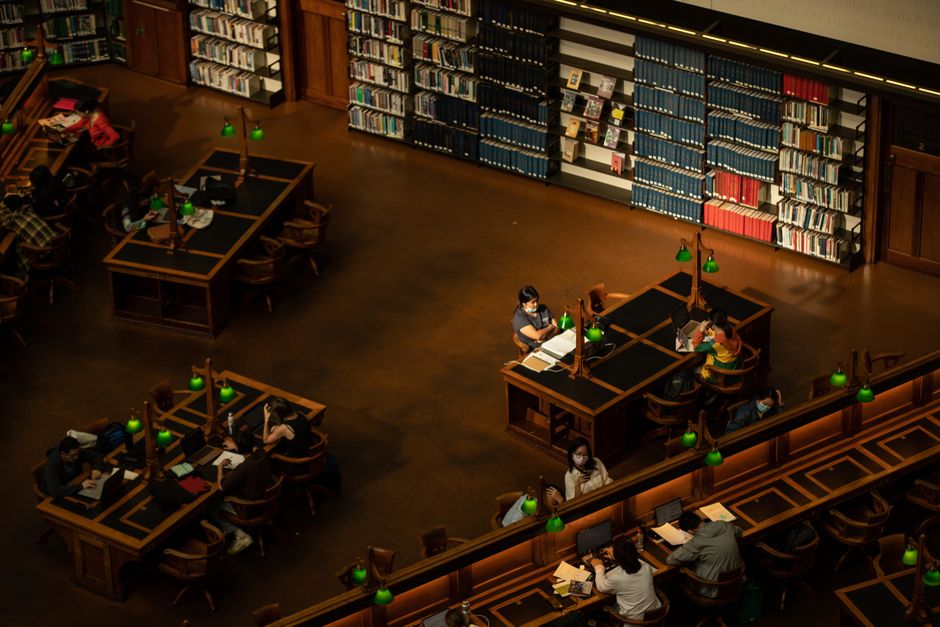Estudantes estudando em uma biblioteca silenciosa com estantes de livros ao fundo, em um ambiente de aprendizagem acadêmica.