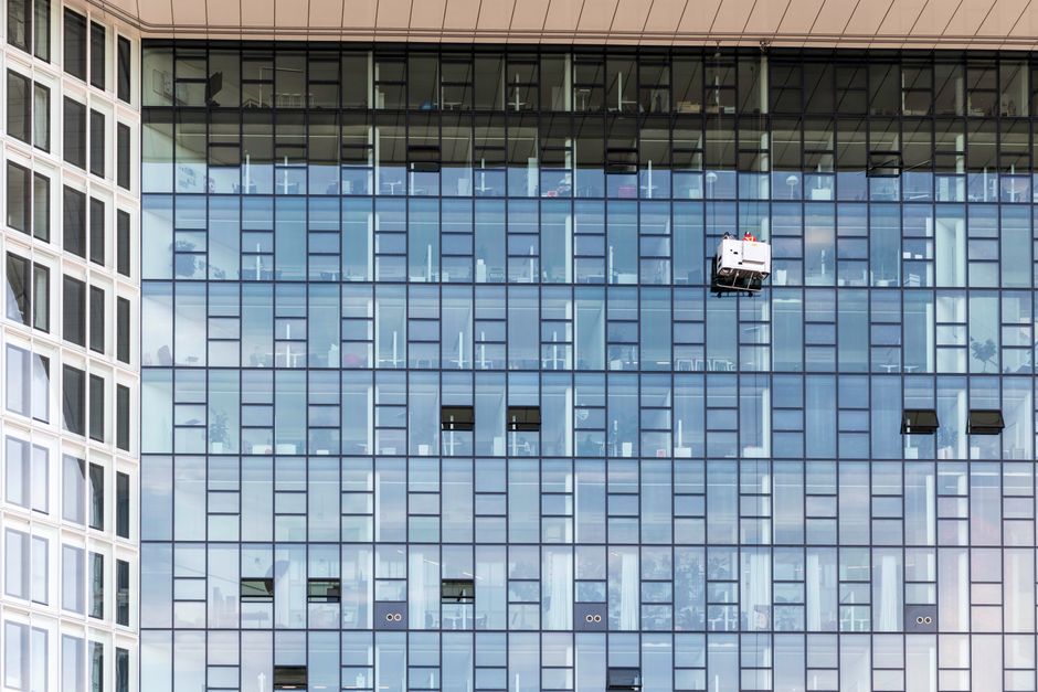 Imagem de um edifício comercial de vidro em construção ou limpeza, refletindo o céu e nuvens.