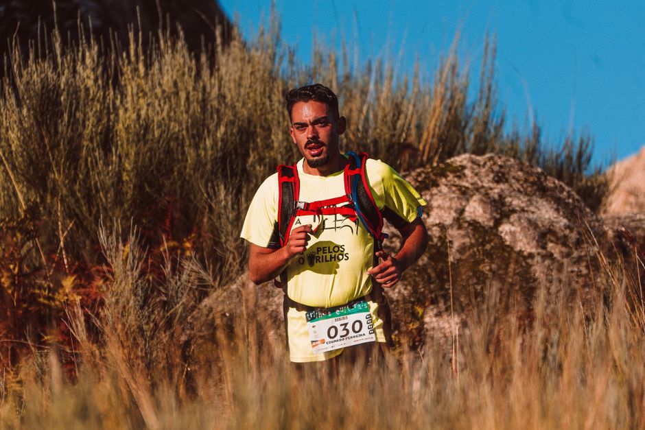 Homem participando de uma corrida de aventura em um ambiente natural ao ar livre, com vegetação e céu azul ao fundo.