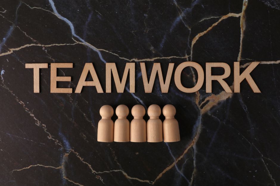 Imagem com a palavra 'TEAMWORK' destacada e figuras humanas de madeira simbolizando cooperação e trabalho em equipe.