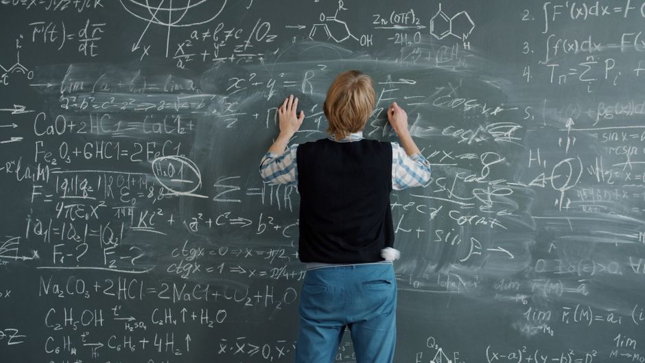 Estudante explicando conceitos complexos de matemática e ciências em uma lousa cheia de fórmulas e anotações, enfatizando o ensino e o aprendizado em sala de aula.