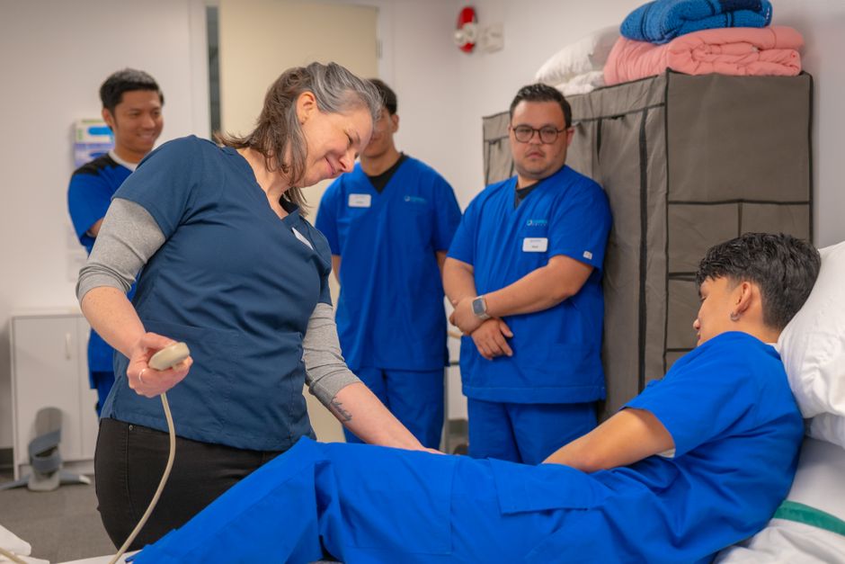Profissional de saúde realizando procedimento em um paciente em ambiente hospitalar, acompanhado por colegas de equipe ao fundo.