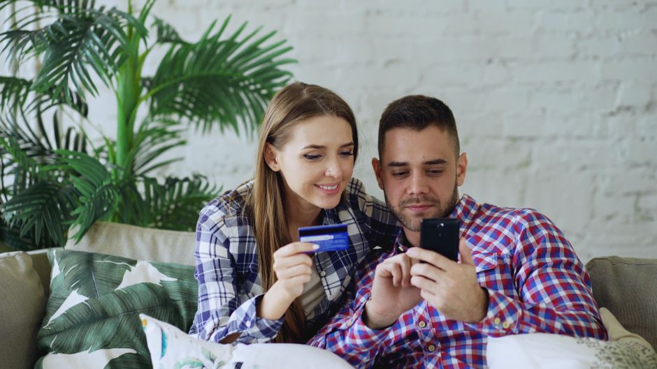 Casal sorridente usando smartphones e cartão de crédito, demonstrando temas de finanças pessoais e consumo digital em um ambiente descontraído.