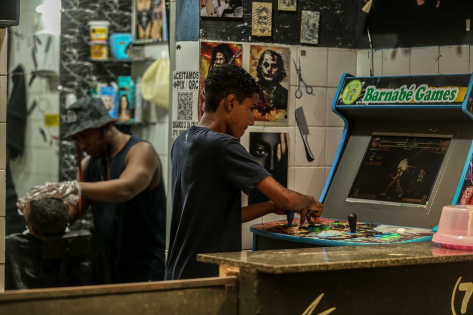 Juvenil jogando arcade em um ambiente de lazer, com decoração e retratos na parede ao fundo.