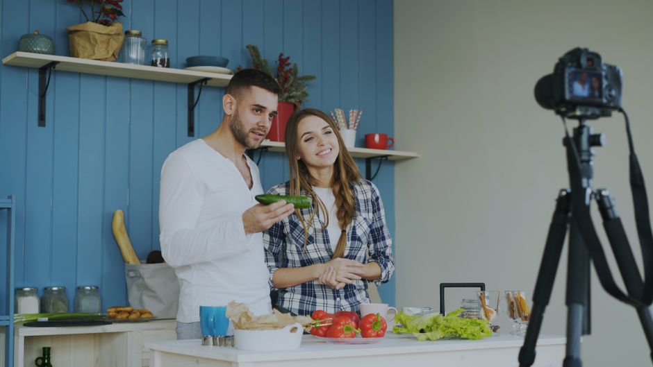 Um casal gravando um vídeo de culinária em uma cozinha decorada de forma moderna, enquanto preparam ingredientes para uma receita.