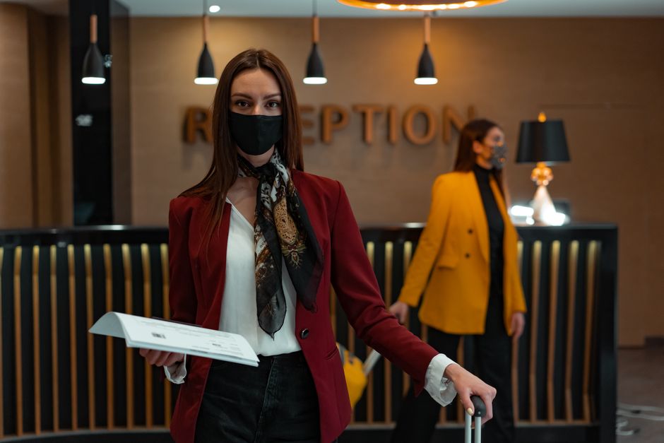 Recepcionista em um hotel ou escritório aguardando visitantes na área de recepção, com atendimento cortês e uso de máscara devido à COVID-19.