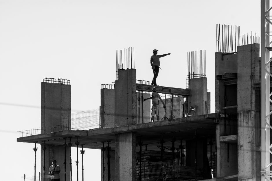 Imagem de construção civil mostrando trabalhadores em uma obra em andamento, destacando o estágio inicial de um edifício com pilares de concreto e reforço de ferro.