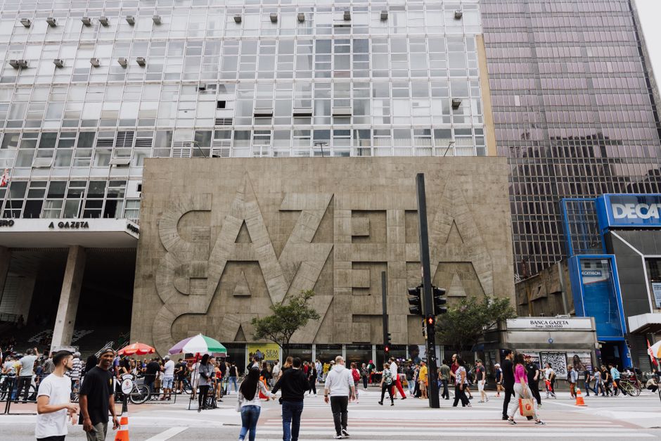 Imagem de uma movimentada área urbana com edifícios comerciais, pessoas atravessando a rua e uma grande fachada com inscrição artística, ilustrando o cenário de uma cidade grande em movimento.