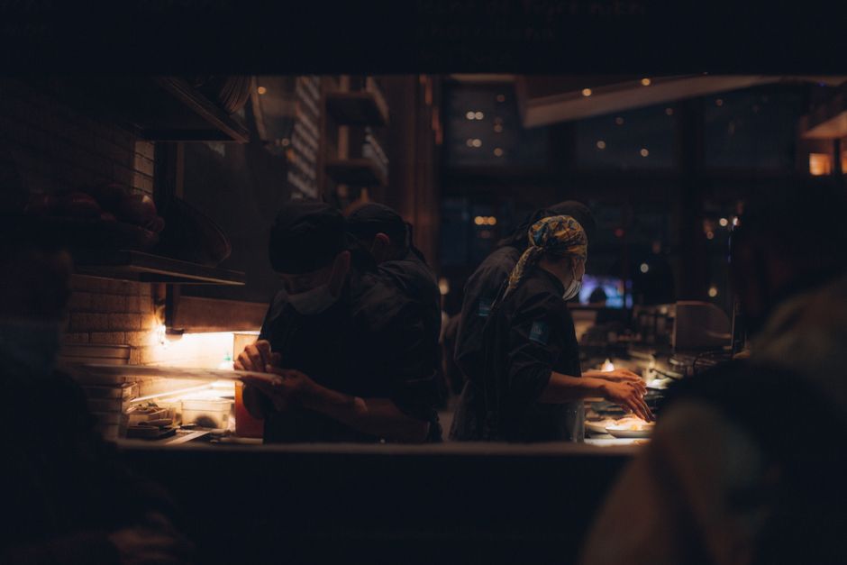 Funcionários de cozinha preparando pratos em um ambiente de restaurante à noite, com iluminação aconchegante e atmosfera movimentada.