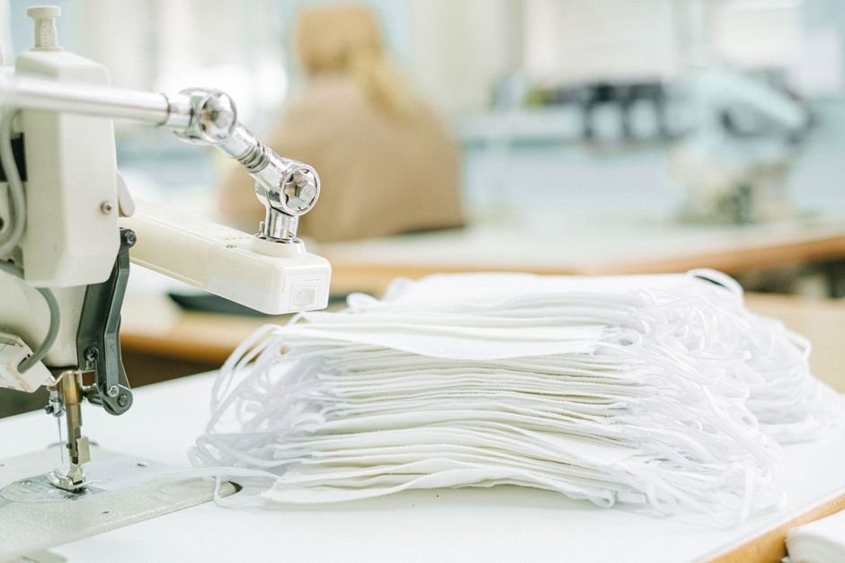 Máquina de costura ao lado de pilhas de máscaras de tecido, simbolizando produção de Equipamentos de Proteção Individual (EPIs) em um ambiente industrial ou de costura de máscara facial.