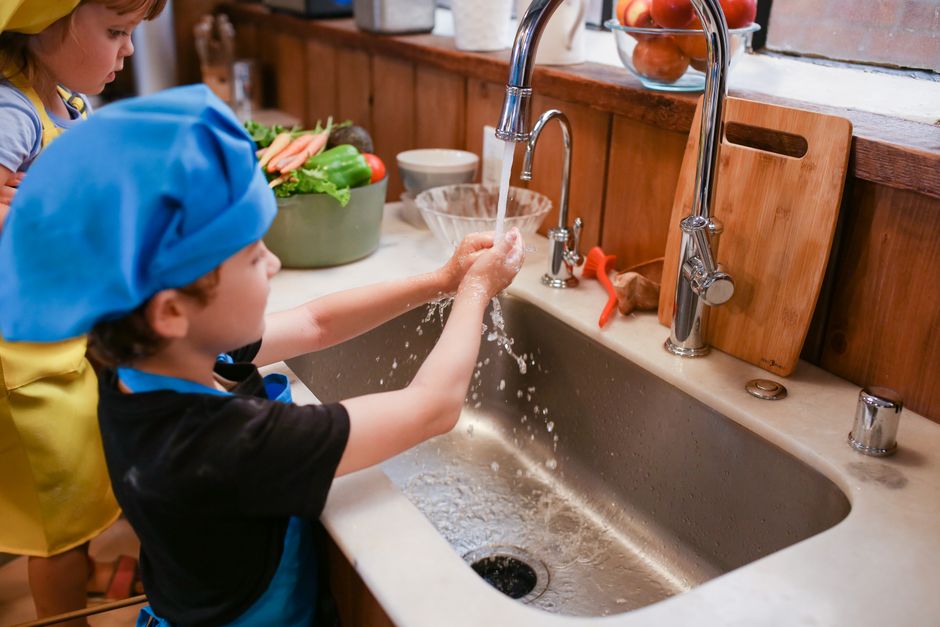 Crianças aprendendo a lavar as mãos na cozinha, promovendo higiene e desenvolvimento de habilidades domésticas.