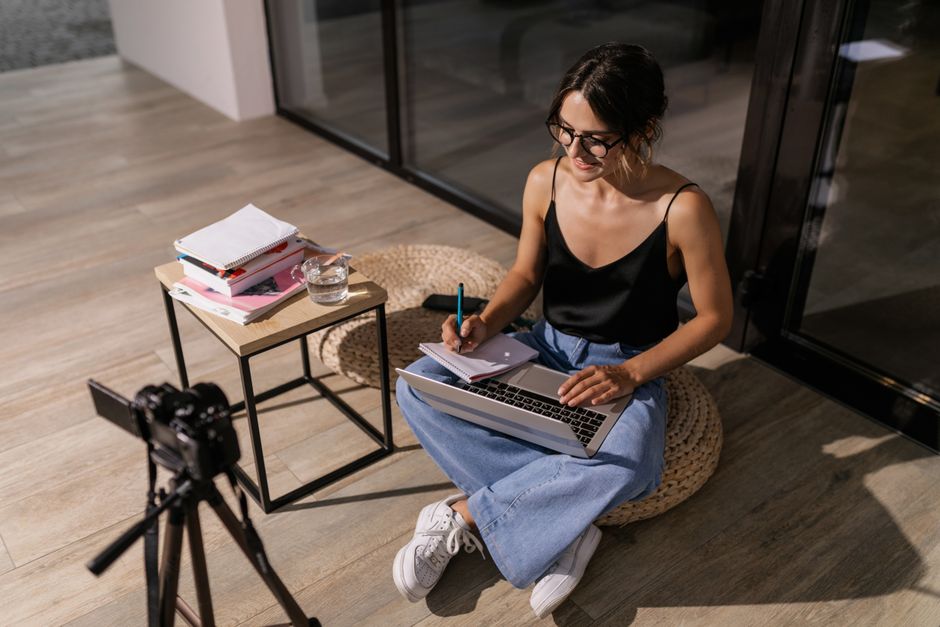 Jovem trabalhando de forma descontraída com computador e caderno, criando conteúdo para um blog de estilo de vida ou produtividade, em um ambiente moderno e confortável.