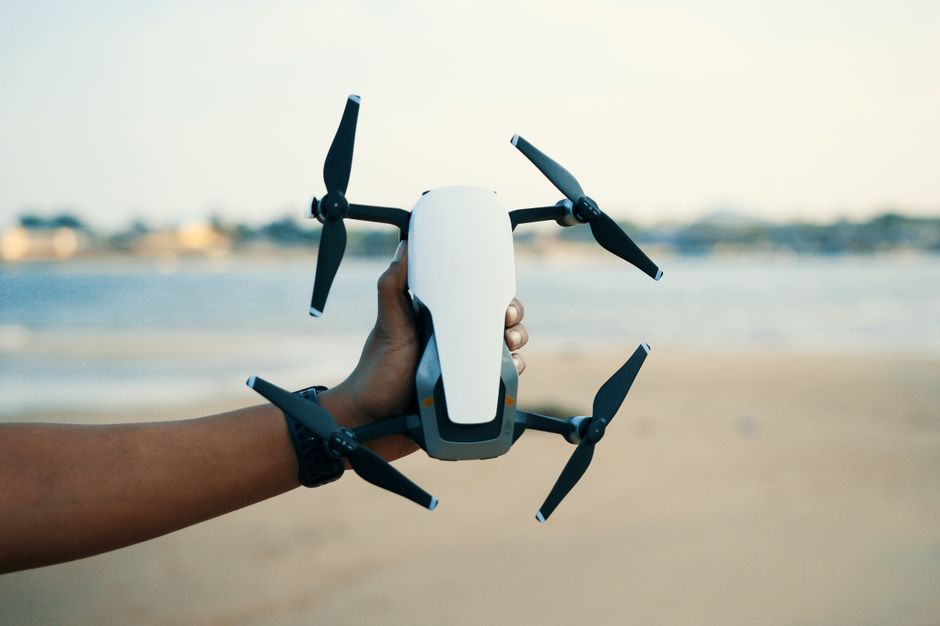 Pessoa segurando um drone com câmeras, pronto para voo ao ar livre, em um cenário de praia ao entardecer.
