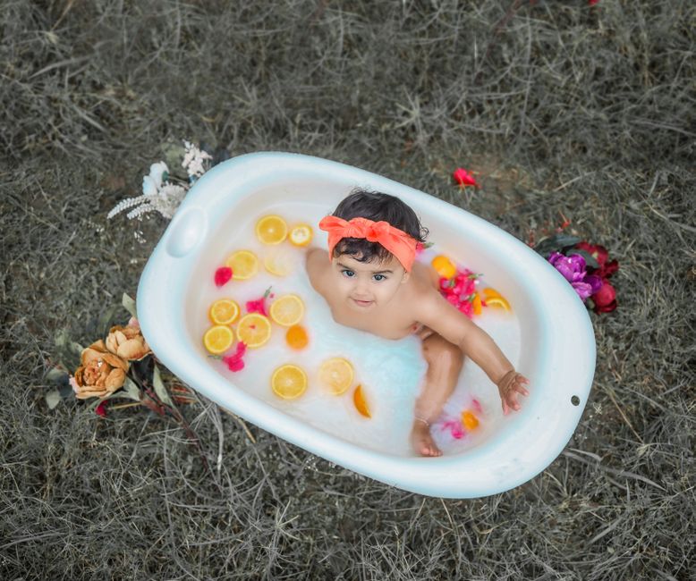 Menina brincando de banho de bolo na área externa, com decoração floral ao redor e elementos coloridos na banheira.