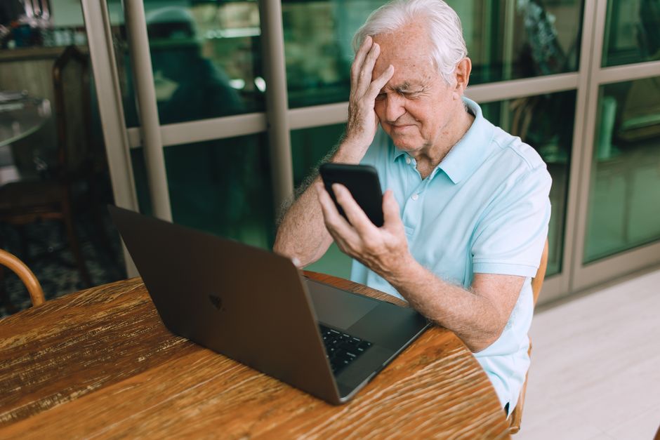 Idoso preocupado usando smartphone enquanto trabalha em frente ao computador, ilustrando questões relacionadas à tecnologia e envelhecimento ativo.