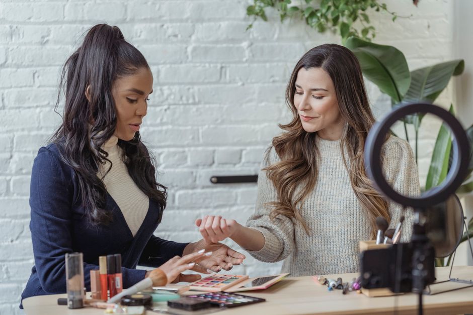 Duas mulheres realizando uma sessão de maquiagem ou consultoria de beleza em um ambiente bem iluminado e decorado com plantas, ideal para um blog de beleza ou bem-estar.