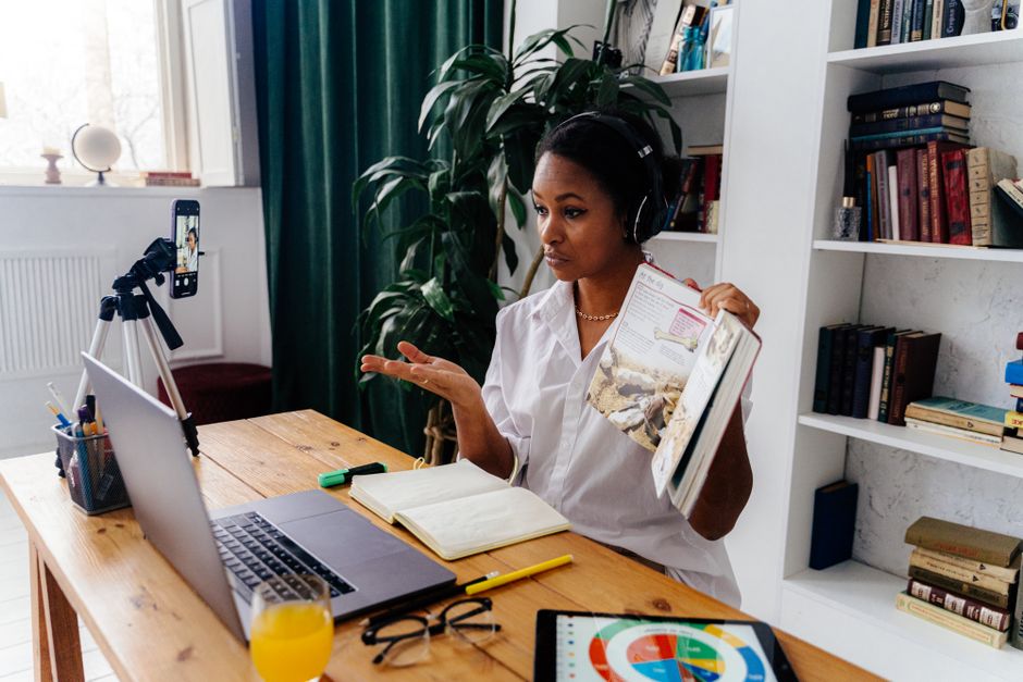 Mulher estudando ou apresentando conteúdo em uma videochamada com livros, computador e ambiente de estudo ou trabalho em casa.
