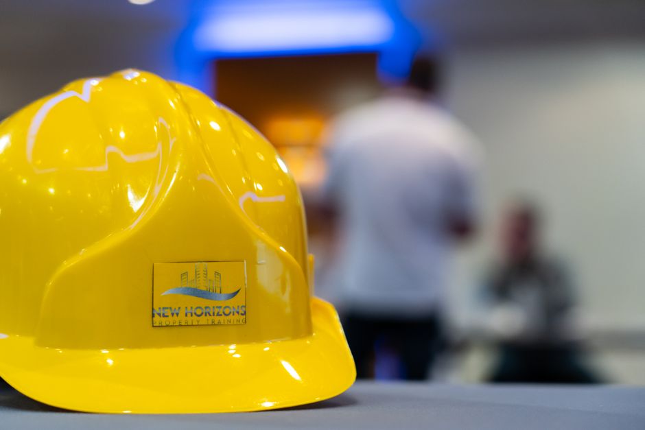 Chapéu de segurança amarelo com o logo de treinamento de segurança, em foco, com profissionais ao fundo em ambiente de treinamento ou construção.