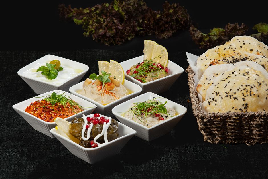 Diversas porções de sobremesas e salgados tradicionais servidos em pequenos recipientes de porcelana na mesa, acompanhados de pão sírio ou árabe, representando a culinária do Oriente Médio ou mediterrânea.