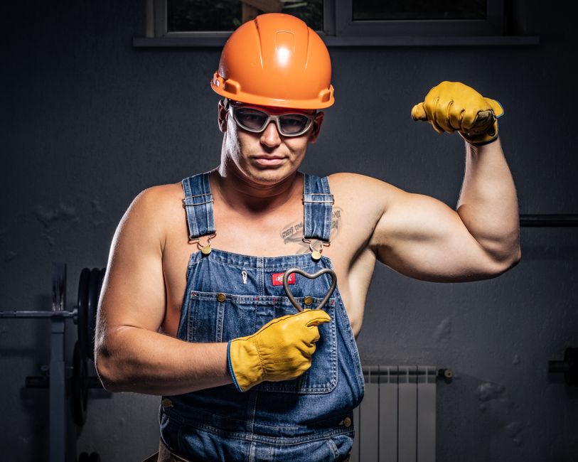 Homem com aparência forte e confiante vestindo roupa de trabalho, usando capacete de segurança e luvas, em ambiente de construção ou manutenção.