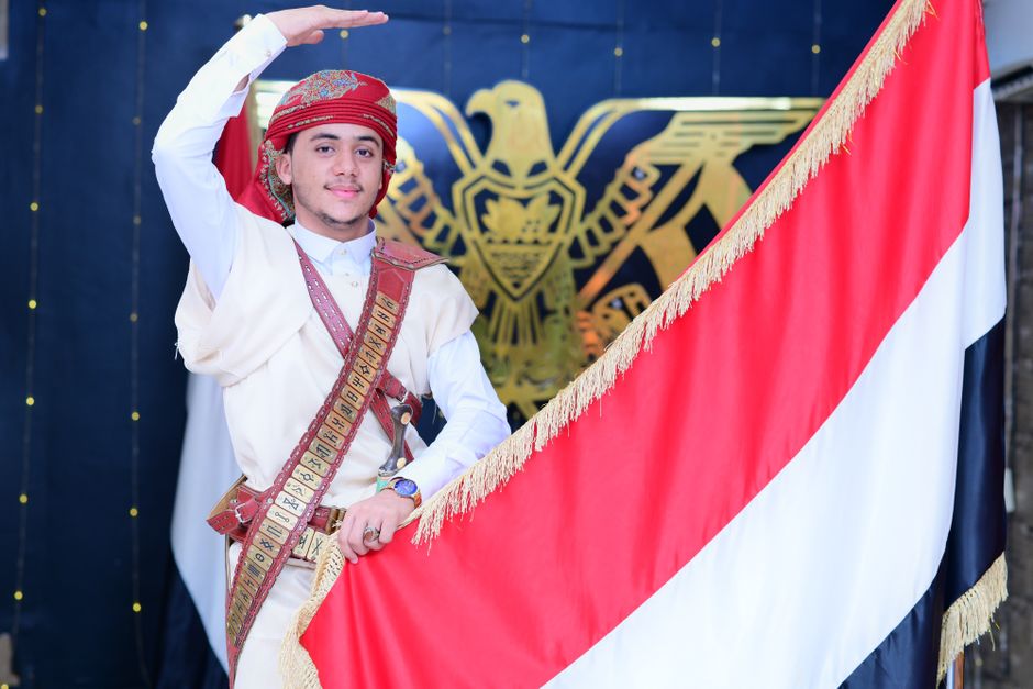 Jovem em traje tradicional fazendo o gesto de honra ao lado de uma bandeira nacional, simbolizando patriotismo e cultura regional.