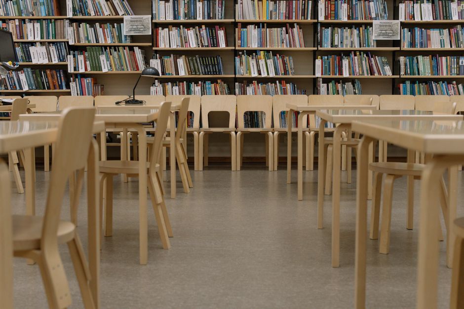 Salão de biblioteca ou espaço de leitura com diversas estantes cheias de livros e cadeiras de madeira ao centro.