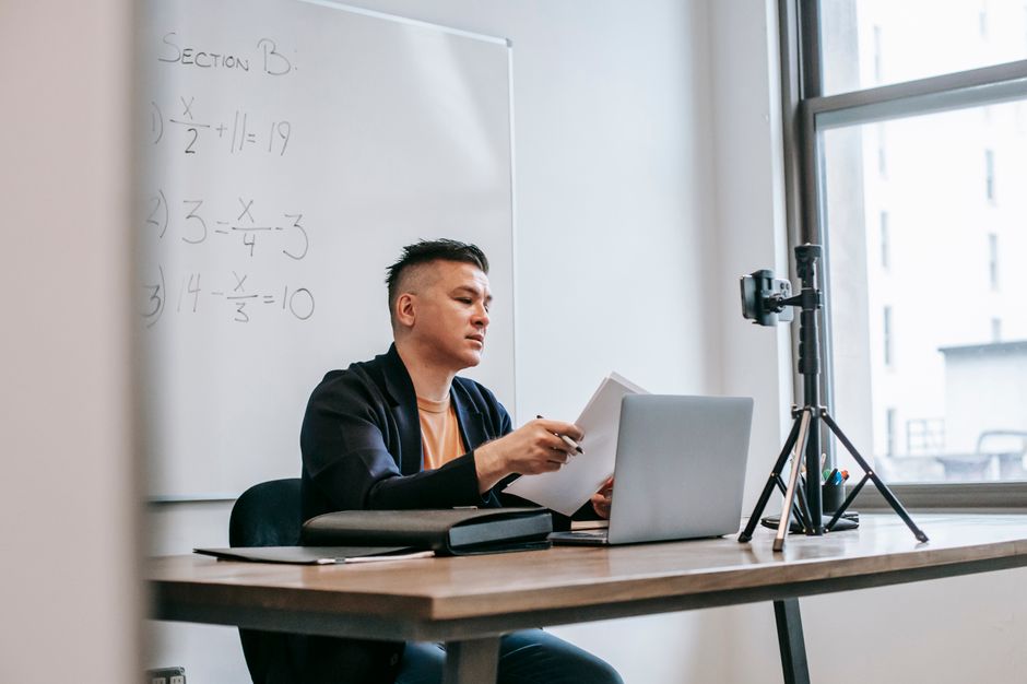 Homem estudando matemática e preparando conteúdo para aula, com câmera e quadro branco ao fundo em ambiente de sala de aula ou home office.